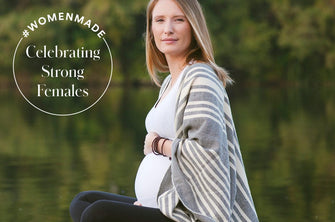Allie Lindenmuth, founder of Anook Athletics, poses in front of a glassy lake surrounding by trees while pregnant and wearing a light gray striped cardigan.