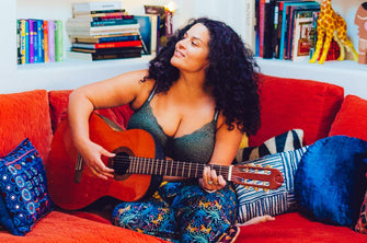 Justina Bdlakeney happily strumming her guitar on a red-orange couch while wearing ThirdLove's Artisan Lace Balconette Bra in Fern.